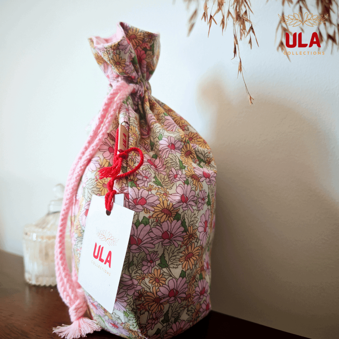 A side profile of the bag standing on a dark wooden ledge. A white "ULA Collections" brand tag is tied to the side with a red string.