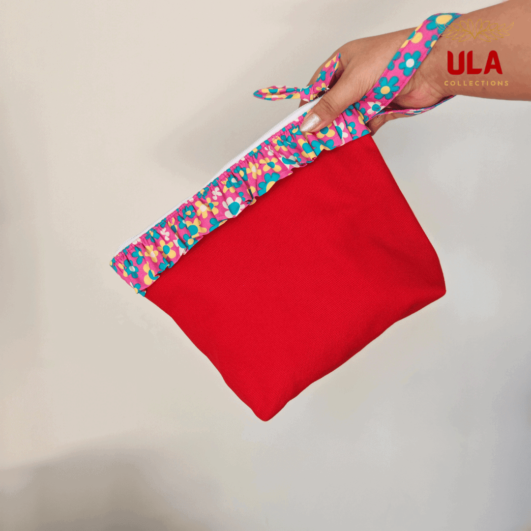 A close-up photograph of a vibrant red canvas pouch being held by a hand. The top of the bag features a colorful, floral ruffle trim in shades of pink, yellow, and turquoise. A matching patterned wristlet strap is looped around the person's wrist. The pouch has a white zipper closure at the top, and the "ULA COLLECTIONS" logo is visible in the upper right corner.