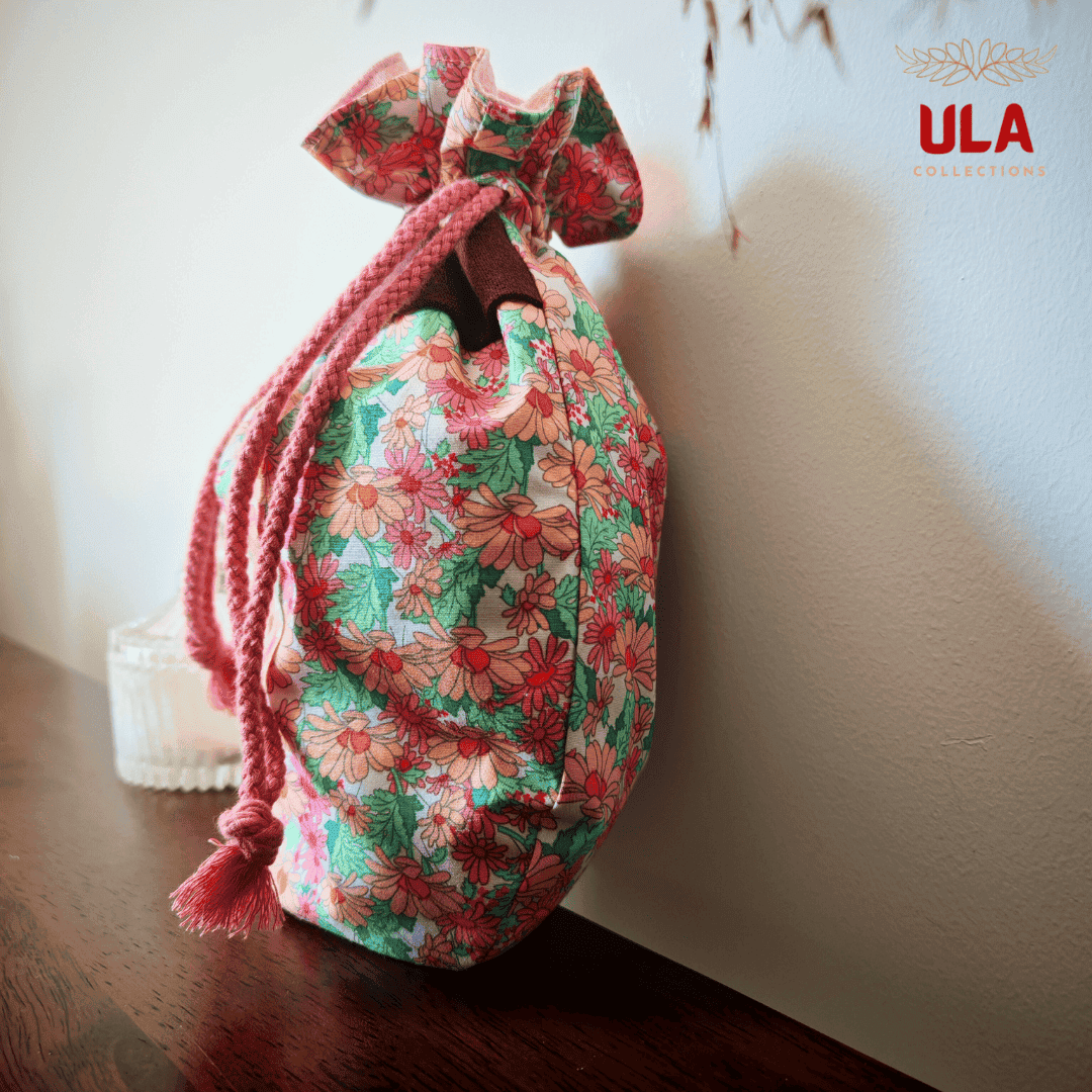 A side profile of the "Sassy" bag sitting on a dark wooden surface against a white wall. Dried wheat stalks hang from the top of the frame, and the ULA Collections logo is in the top right corner.