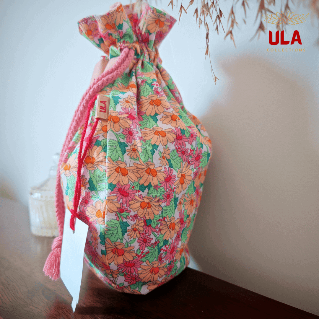 A side-angle view of a rounded, drawstring fabric bag sitting on a dark wooden surface. The bag features a vibrant floral pattern with orange, pink, and red daisies on a light background with green leaves. It has a thick, pink braided drawstring and a small leather brand tag that reads "ULA."