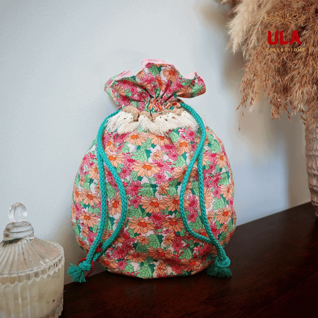 A full front-facing view of a rounded fabric drawstring bag sitting on a dark wooden surface. The bag features a dense floral print of orange, pink, and red daisies with green leaves. It is cinched at the top, accented by a row of cream-colored fringe tassels and thick teal braided ropes that hang down the sides.