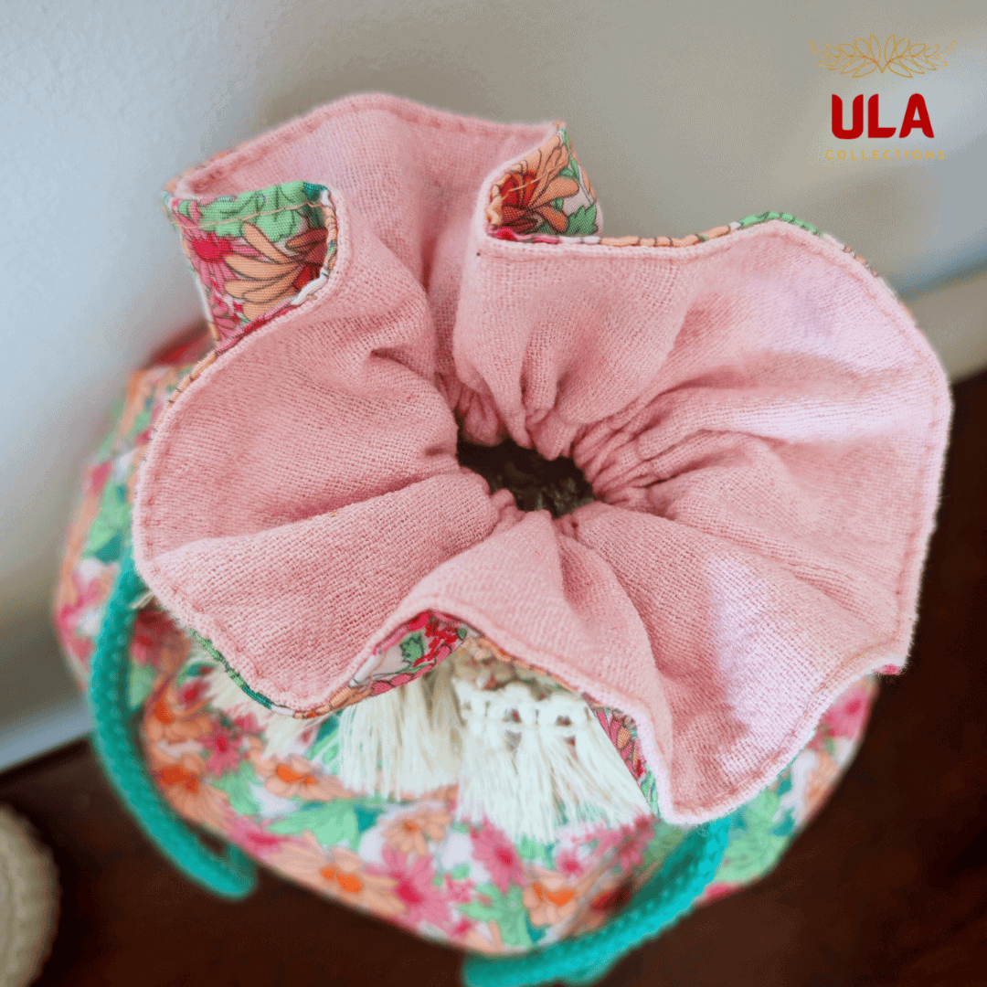 A top-down view looking into the opened drawstring bag. The interior is lined with a soft, plush pink fabric that ruffles around the circular opening. White decorative tassels and teal braided drawstrings are partially visible against the floral exterior.