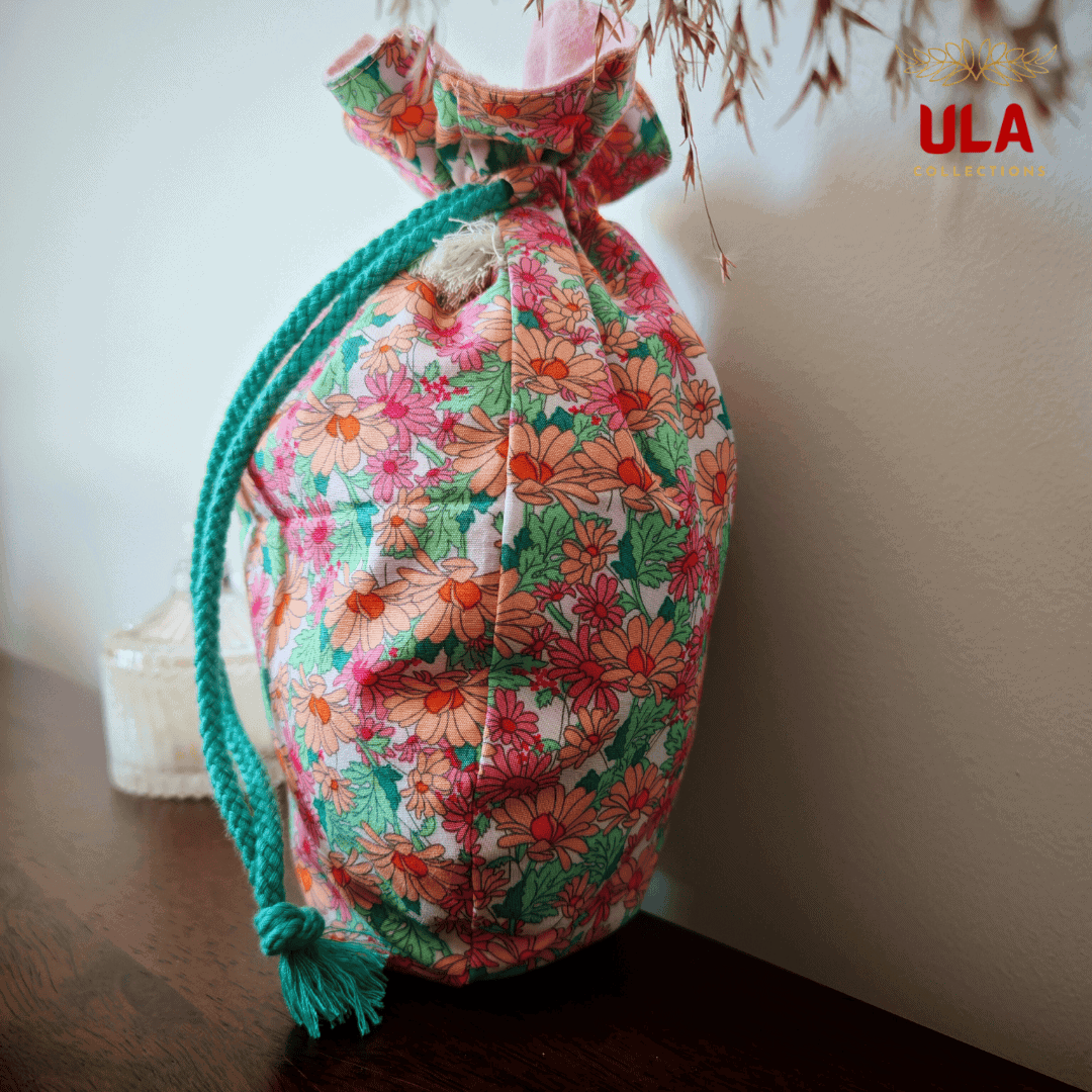 A side-angle view of the floral drawstring bag, showcasing its plump, upright shape. The teal braided drawstring is draped over the side, ending in a knotted tassel. The background features a plain light-colored wall and a glass jar on the wooden surface.