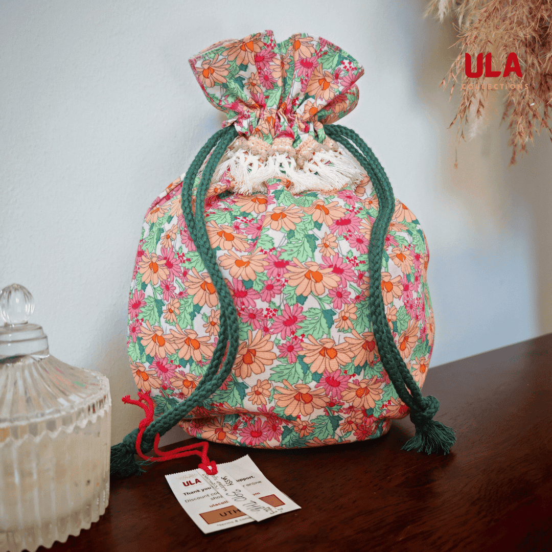 A front view of a similar floral drawstring bag sitting on a wooden surface next to a paper tag. This version features dark forest-green braided drawstrings and a row of cream fringe tassels. The "ULA Collections" logo is visible in the upper right corner.