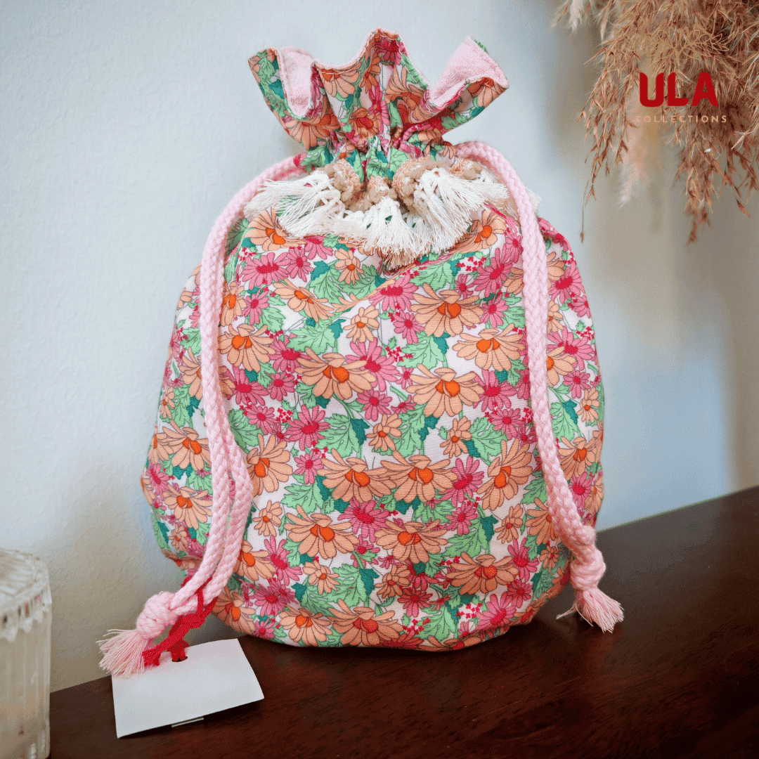 A full front view of the bag with its light pink drawstrings and cream fringe tassels. A white paper tag is attached to the left drawstring.