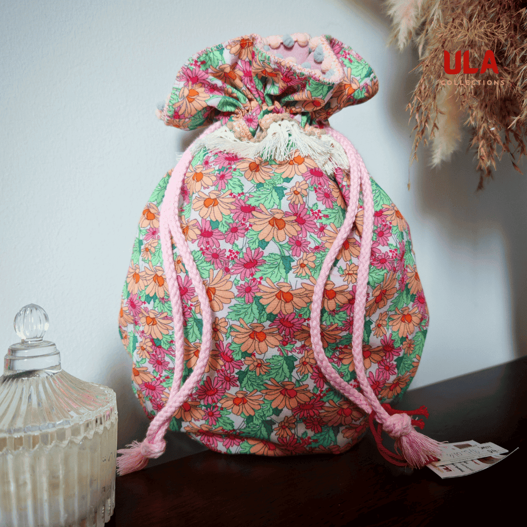 A front-facing view of a rounded, floral drawstring bag sitting on a dark wooden surface. The bag features a vibrant orange, pink, and red daisy print on a light background with green leaves. It is cinched at the top with thick, light pink braided ropes and accented by a row of cream-colored fringe tassels.