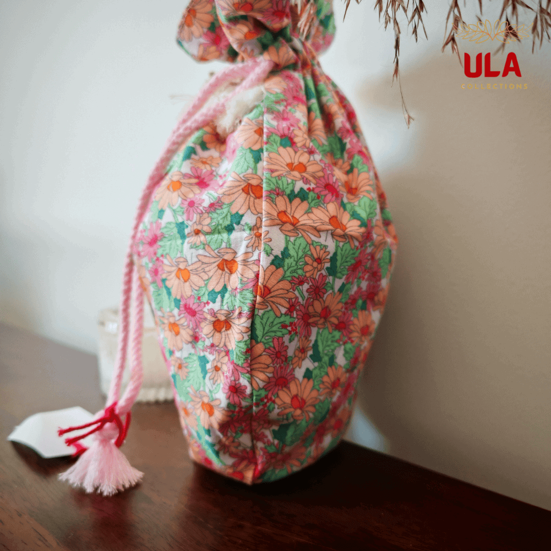 A side view of the pouch, highlighting its plump, gathered shape and the colorful daisy pattern. One of the light pink braided drawstrings hangs down the side, ending in a tassel.