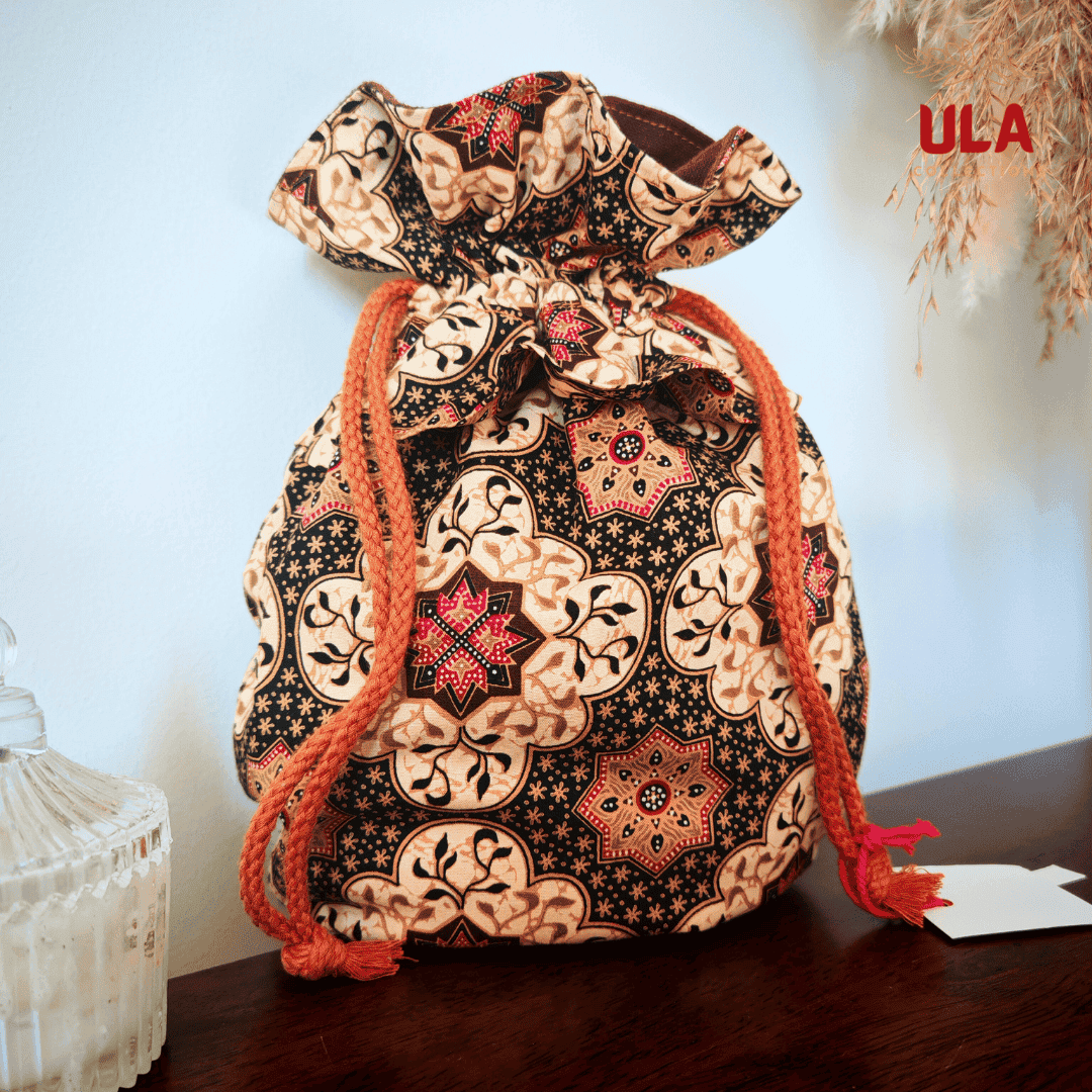 A studio photograph of a cinched drawstring bag made from fabric with an intricate batik-style pattern in cream, black, and reddish-pink. The bag features thick, orange braided drawstrings and is set on a dark wooden surface against a neutral wall. Dried wheat-like stalks hang from above, and the ULA Collections logo is in the top right corner.