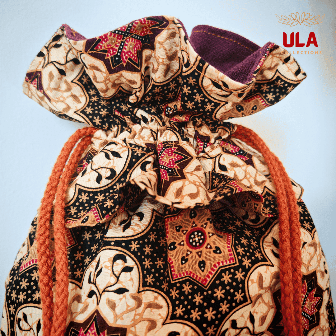 A close-up shot focusing on the upper half of the drawstring bag. The detail highlights the texture of the orange braided cord and the sharp print of the fabric, which includes star-shaped patterns and leaf-like flourishes. The ruffled top shows the transition from the patterned exterior to the solid dark purple fabric lining. The ULA Collections logo is in the top right corner.