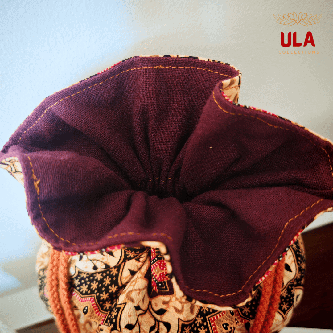 A top-down close-up looking into the open top of the drawstring bag. The focus is on the textured, deep purple or burgundy interior fabric and the clean orange stitching along the top edge. The exterior batik-style pattern and the orange drawstrings are visible at the bottom of the frame. The ULA Collections logo is in the top right corner.