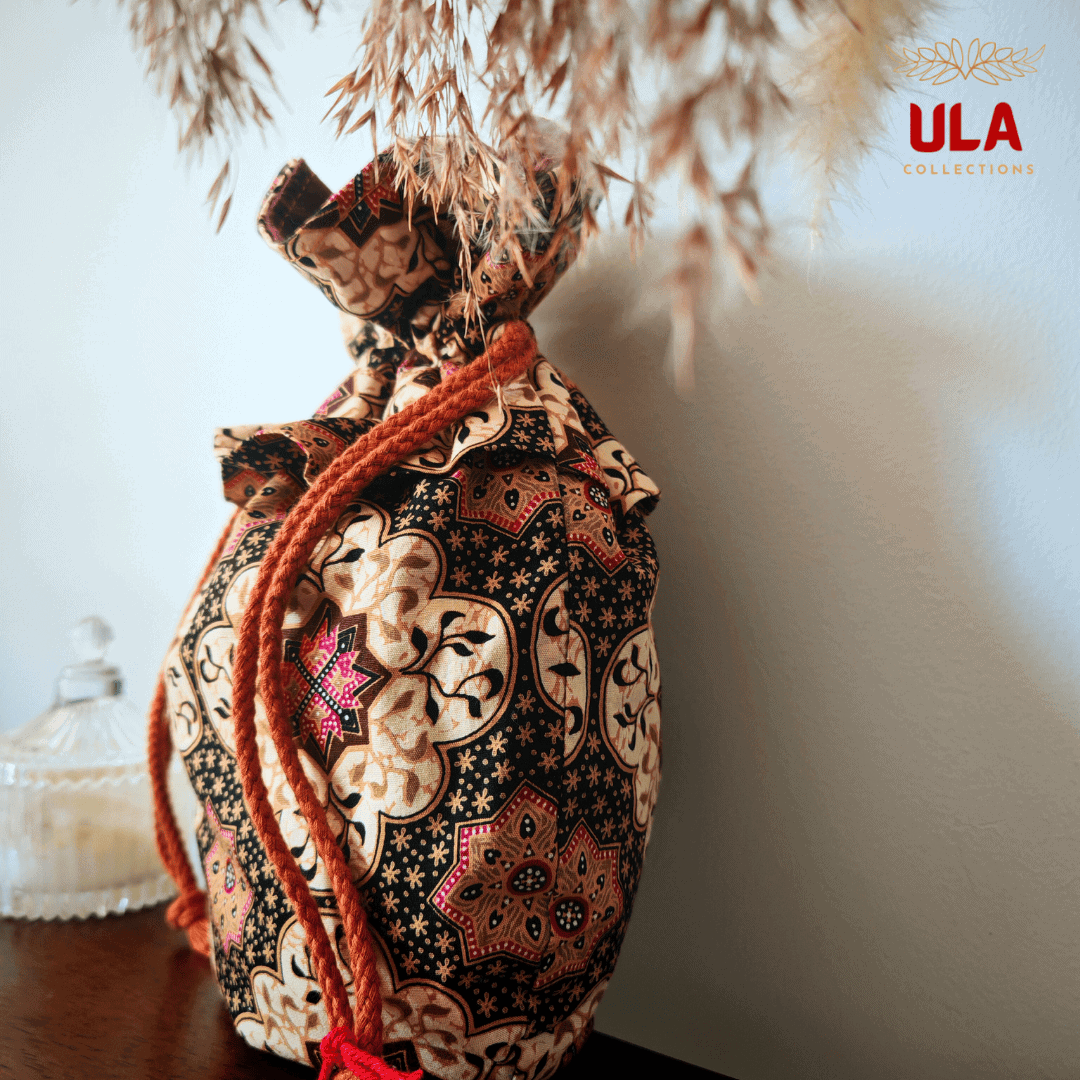 A front-facing view of the drawstring bag sitting on a dark wooden ledge. The pattern features repeating floral and geometric motifs. The top of the bag is ruffled where it is cinched, revealing a glimpse of the deep purple interior lining. To the left, a small decorative glass jar is partially visible. The ULA Collections logo is in the top right corner.