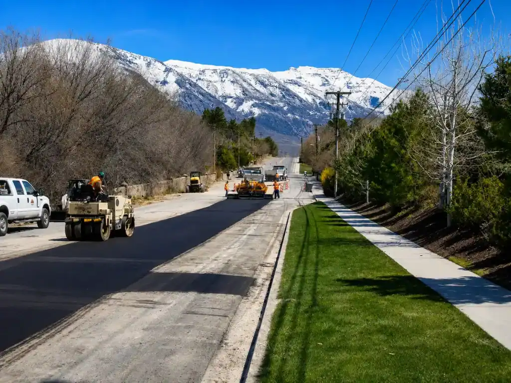 Road paving and asphalt patching project on a Utah roadway with construction equipment and snow-capped mountains in the background.