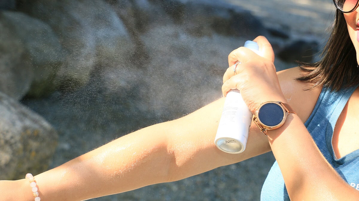 A golfer in a sleeveless top sprays sunscreen onto their arm during a sunny round, ensuring proper coverage by applying and rubbing in the product for an even protective layer.