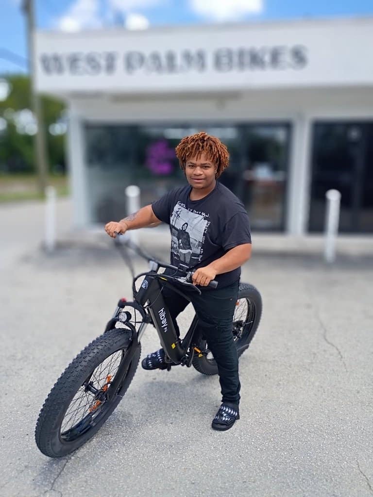 Young boy with curly hair riding a black mountain bike outside West Palm Bikes store.