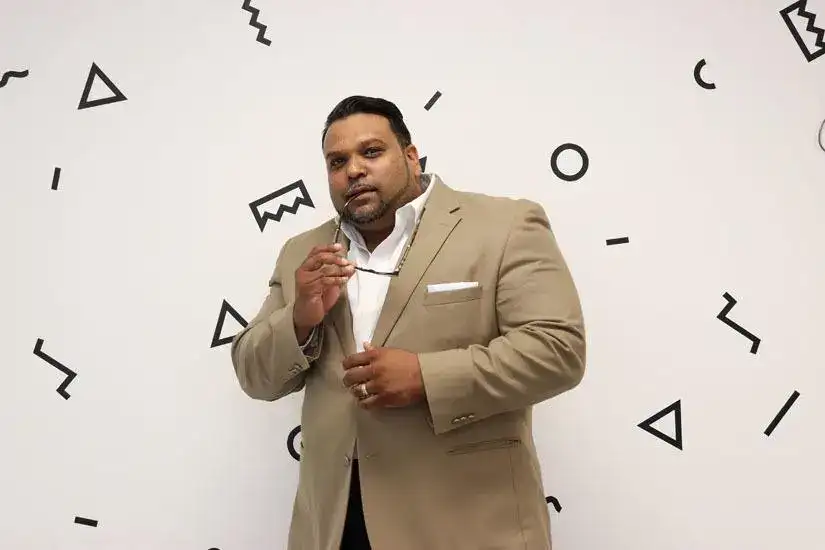 Businessman in beige suit holding glasses against geometric patterned wall.