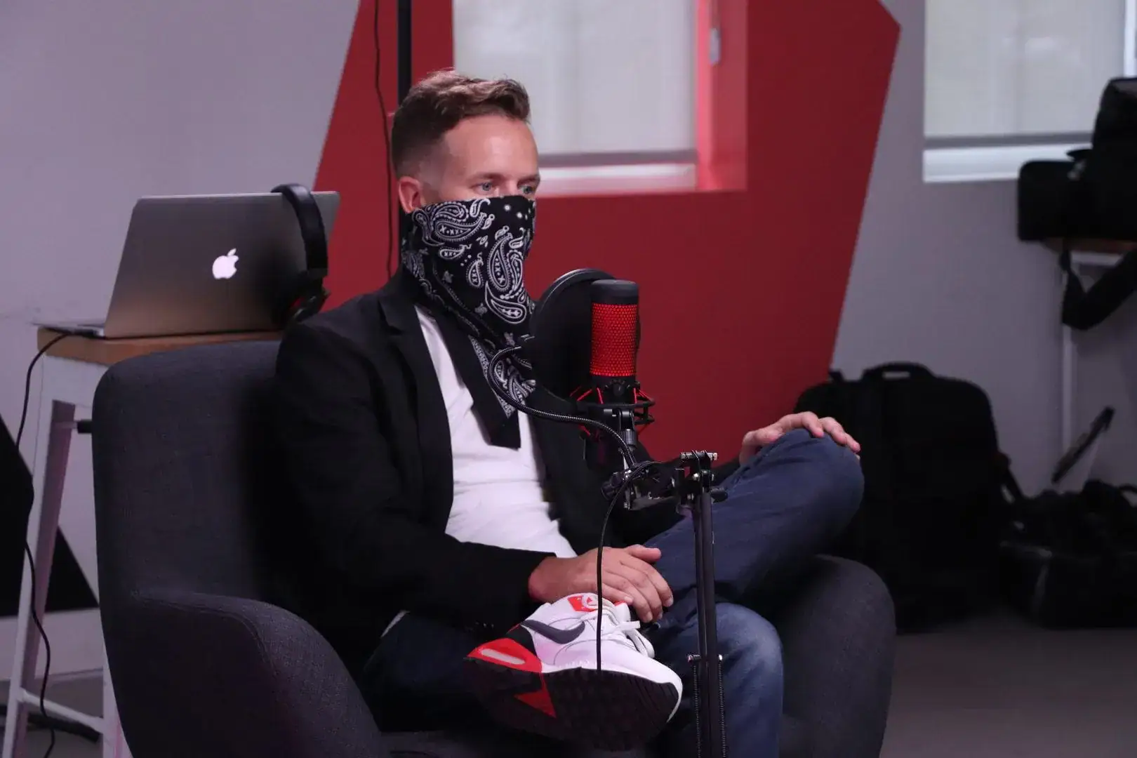 Podcast host wearing a bandana mask speaking into a microphone in a recording studio.