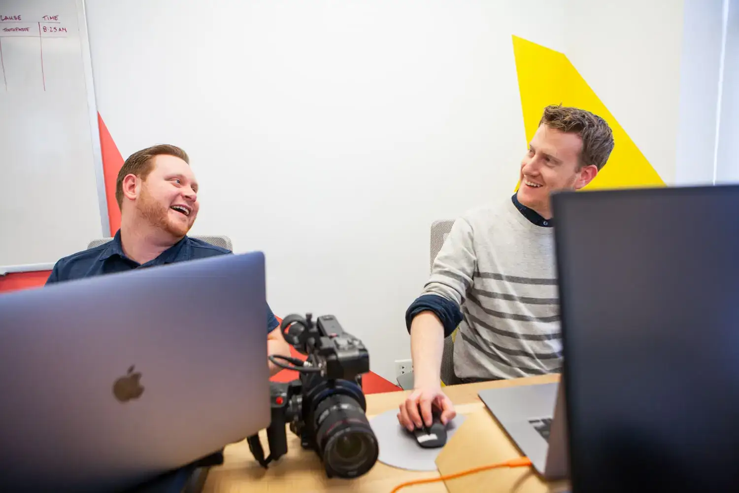 Two men collaborating and smiling at a desk with computers and camera in an office setting.