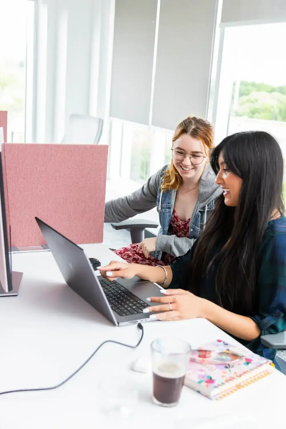 Two women collaborating on a laptop in a bright office setting.