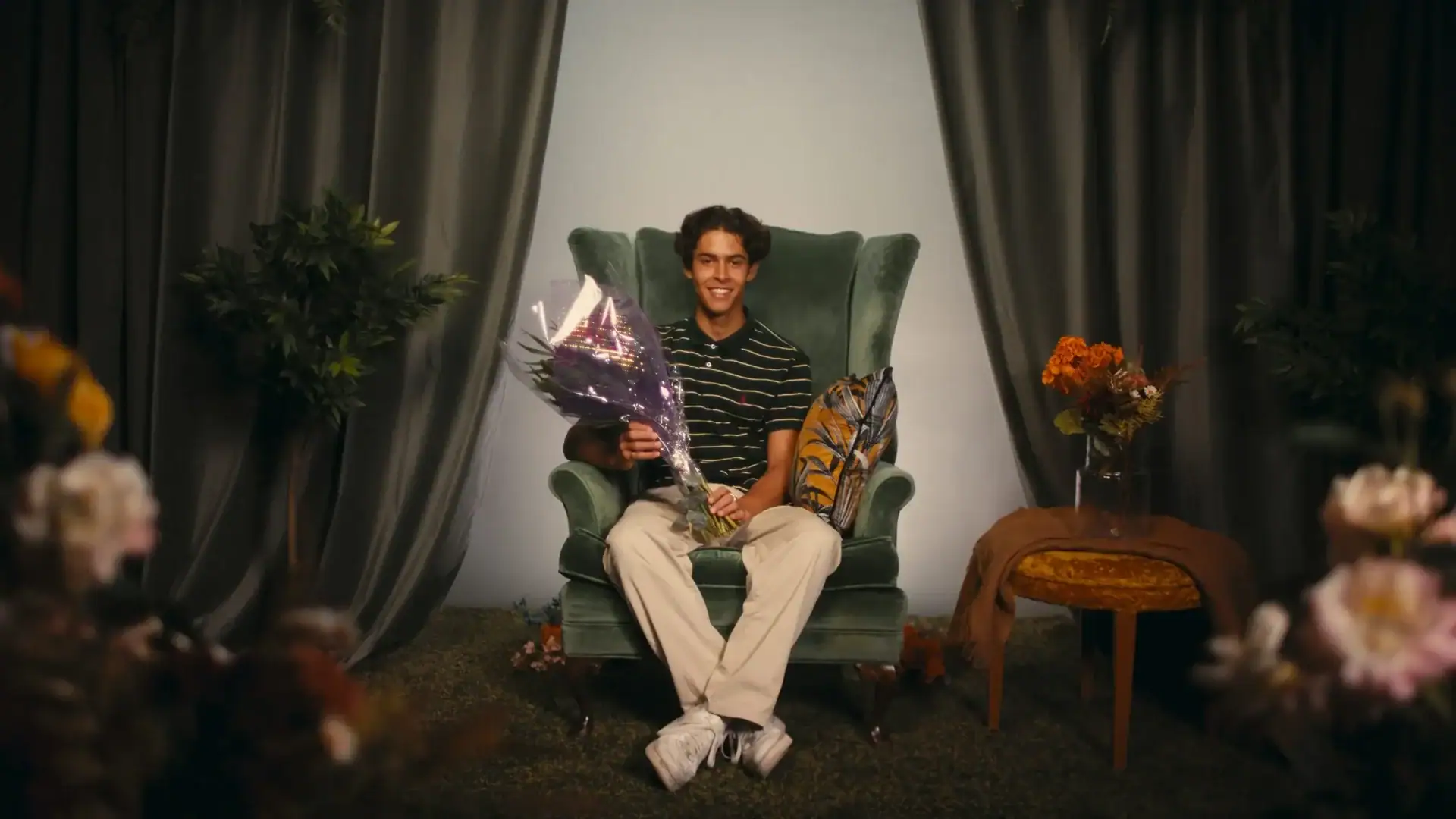Man sitting in a cozy living room with flowers and curtains, smiling and holding a bouquet of flower.