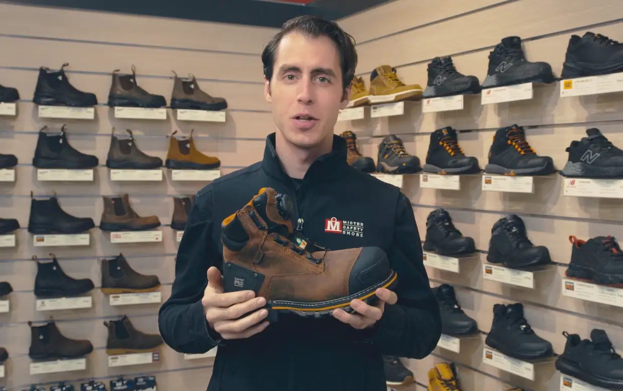 Man showcasing a brown work boot in a shoe store with various footwear on shelves behind him.