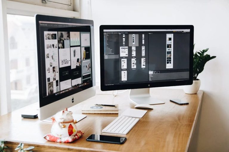 Dual computer screens displaying website and marketing project visuals, on a wooden desk with tech accessories, illustrating authority & marketing technology.