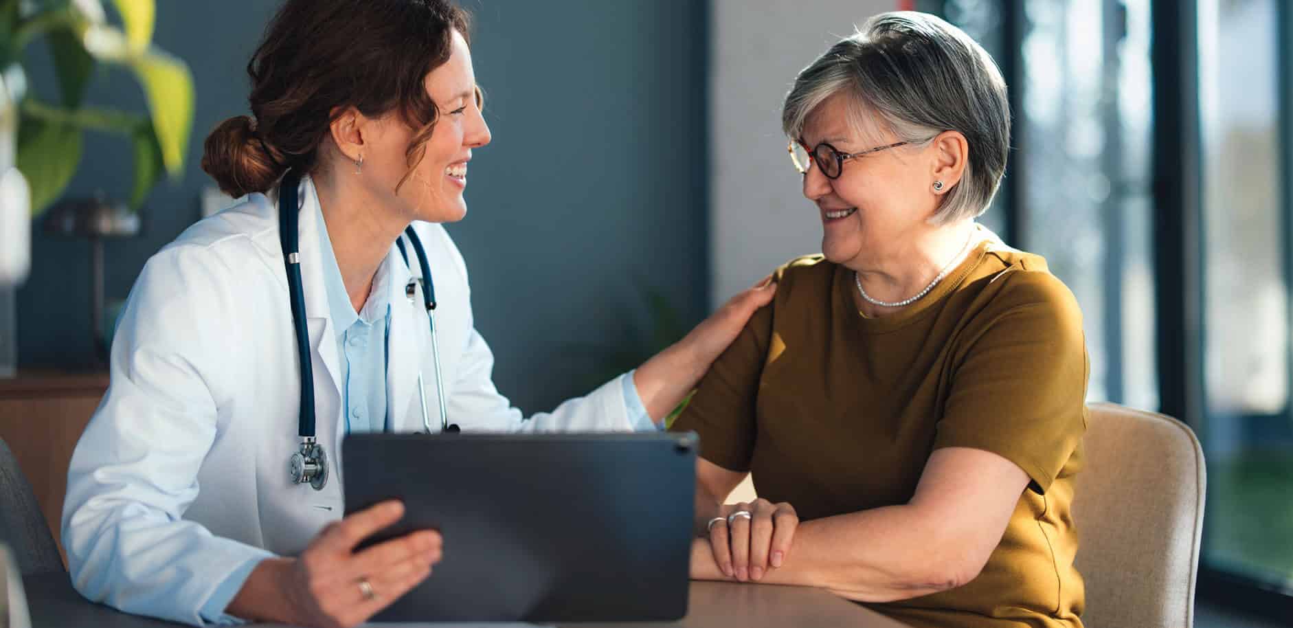 A doctor and senior woman sit together in a bright doctor office reviewing information on a tablet, sharing a trusting medical consultation focused on health