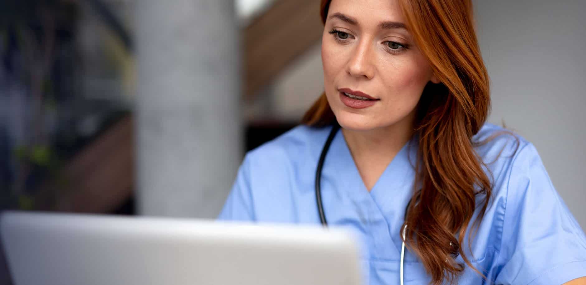 Redhead female doctor in blue scrubs working intently on a laptop, focused on the screen, with a stethoscope draped around her neck, embodying professionalism in healthcare