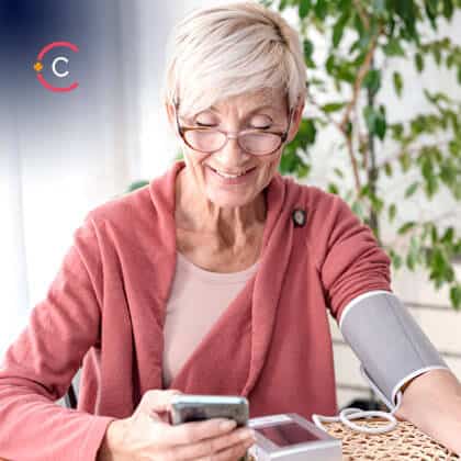 Older woman sitting at her kitchen table, measuring her blood pressure holding a mobile phone in the other hand