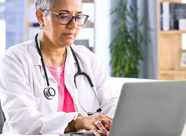 Confident female doctor uses a laptop in her office. She is wearing a lab coat, stethoscope and a pink shirt