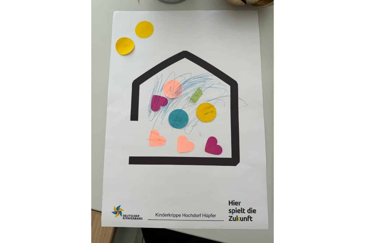 Bunte Kinderzeichnung in Hausform mit Herz- und Kreisaufklebern, symbolisiert Gemeinschaft und Zukun.