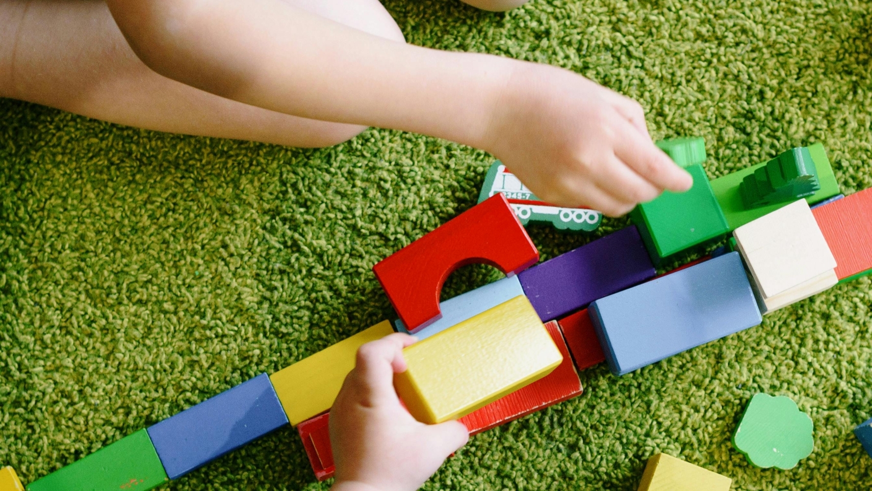 Kinder spielen mit bunten Bauklötzen auf grünem Teppichboden.