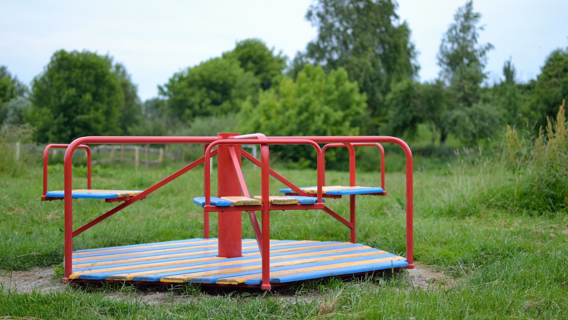 Rotes Karussell auf einem Spielplatz mit grüner Wiese und Bäumen im Hintergrund.