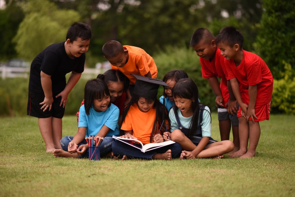 Happy children gathered around a graduate with a cap, reading a book.