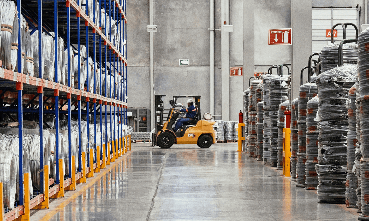 A forklift operator moving pallets in a large warehouse with high shelves filled with goods on the left and right sides. The warehouse has a clean, polished concrete floor and safety signs on the wall.