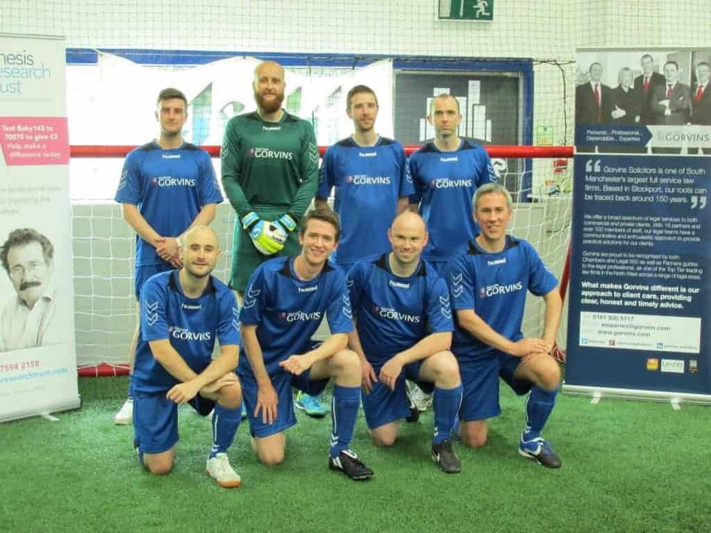 Football team logo wear at indoor sports facility in front of sponsor banners, promoting Gorvins solicitors, legal services in South Manchester, team in blue kits with Gorvins branding.