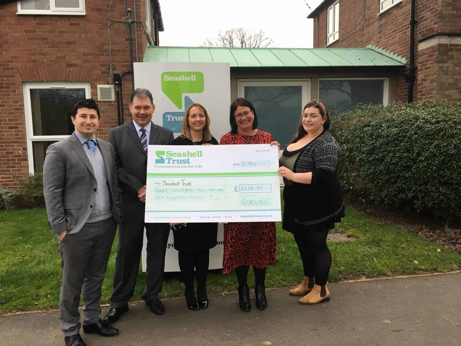 Seashell Trust donation presentation outdoor with five people holding a large cheque, standing in front of a building and greenery.