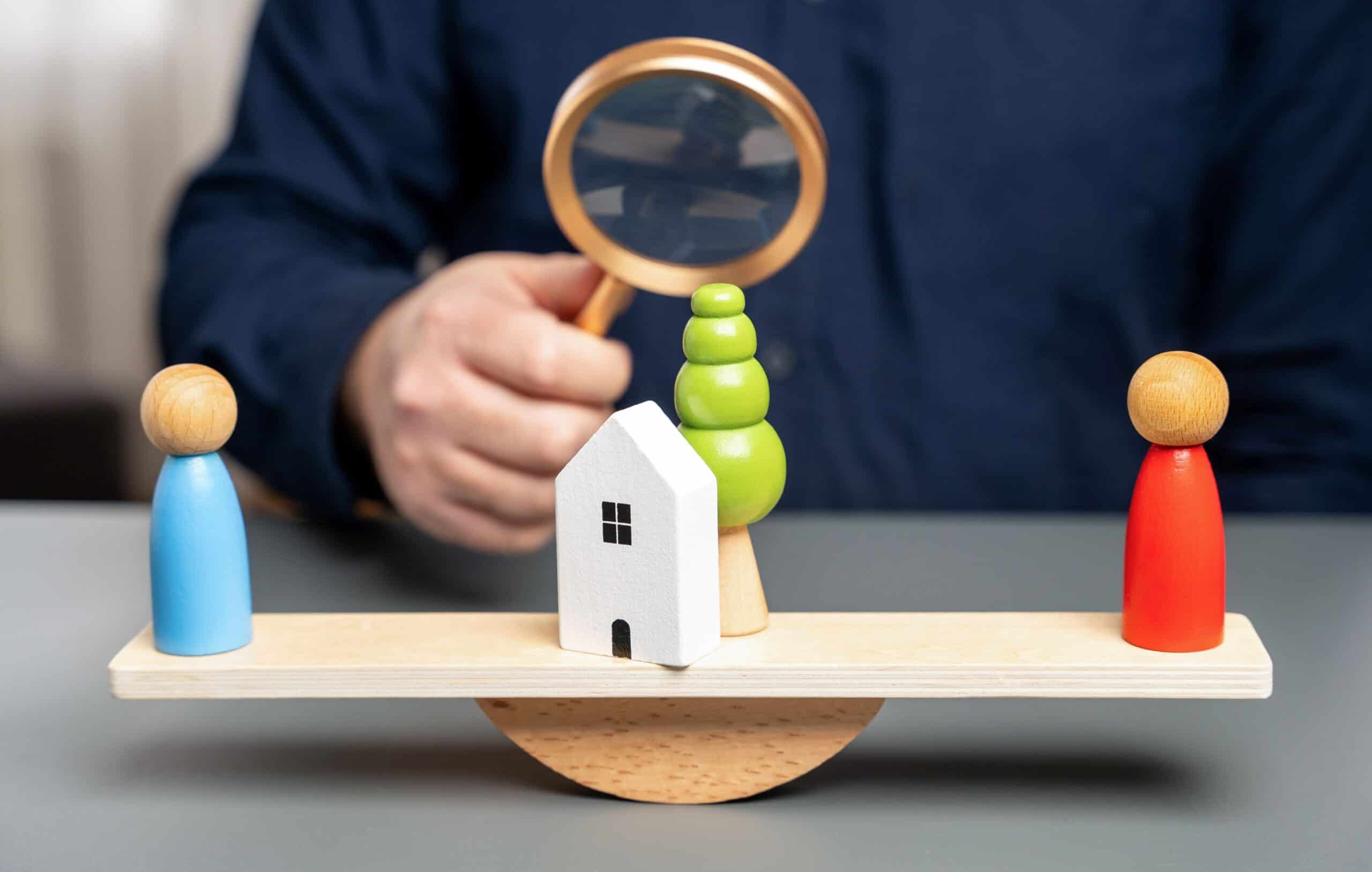 Colourful balanced wooden figurines and a small house model on a wooden seesaw with a person holding a magnifying glass in the background.