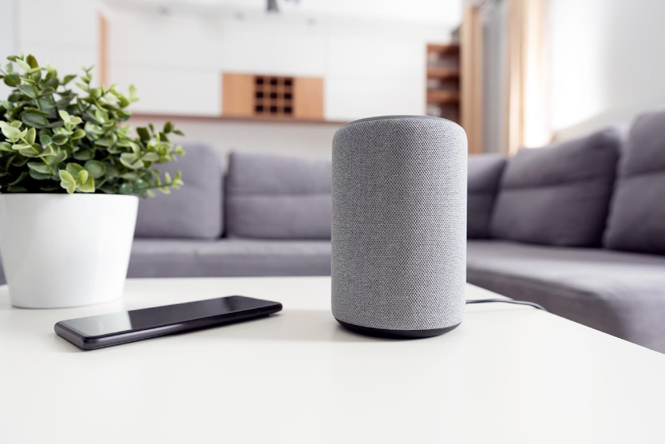 Smart home speaker on a white coffee table with a smartphone and potted plant in a modern living room.