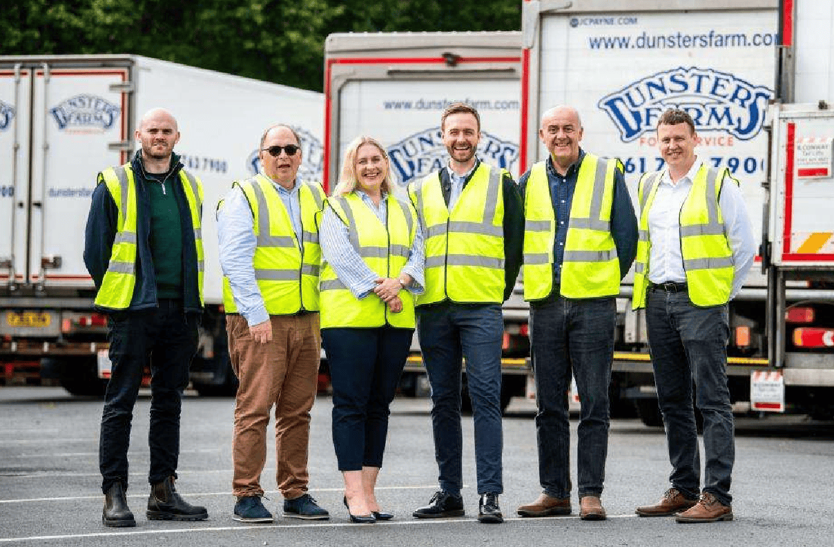 High-visibility team of logistics professionals standing in front of Dunster's Farm delivery trucks during daytime.
