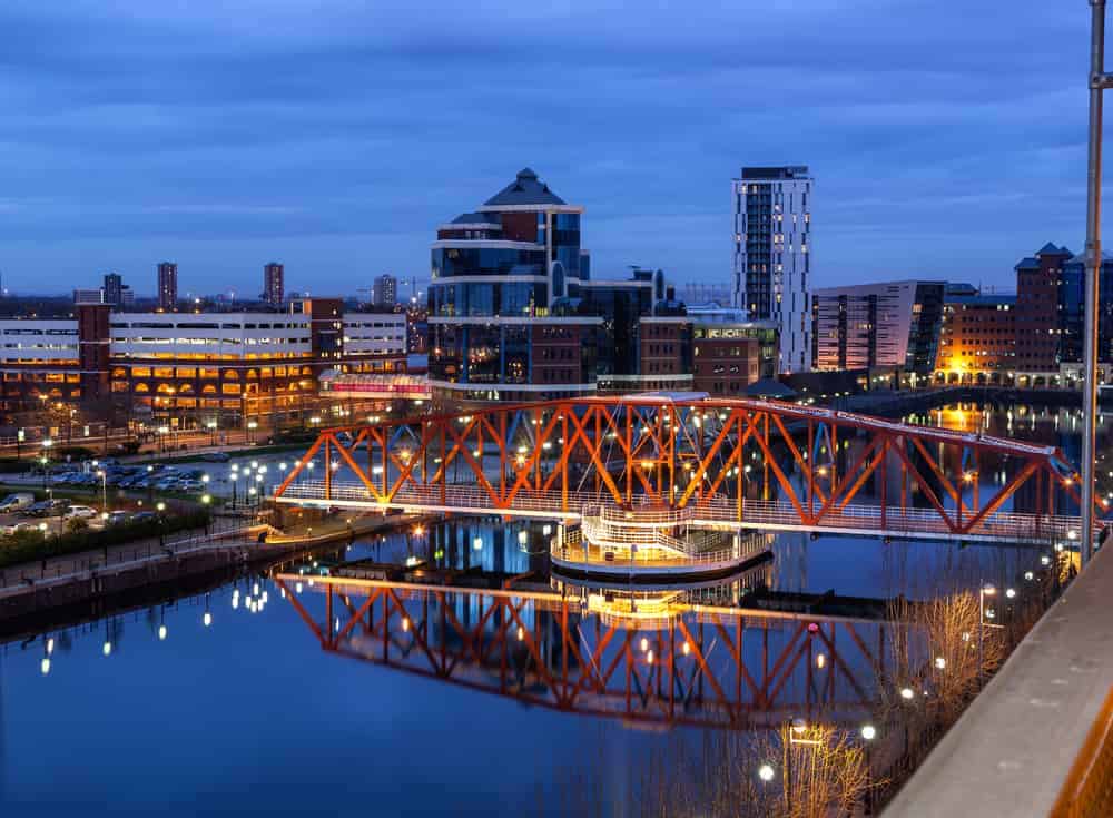 Urban Manchester skyline with illuminated buildings and a bridge over reflective water at dusk.
