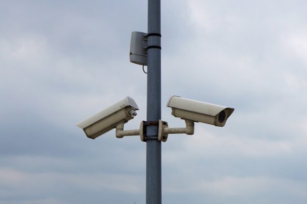 Two white security cameras mounted on a metal pole against a cloudy sky, angled in opposite directions to monitor different areas.
