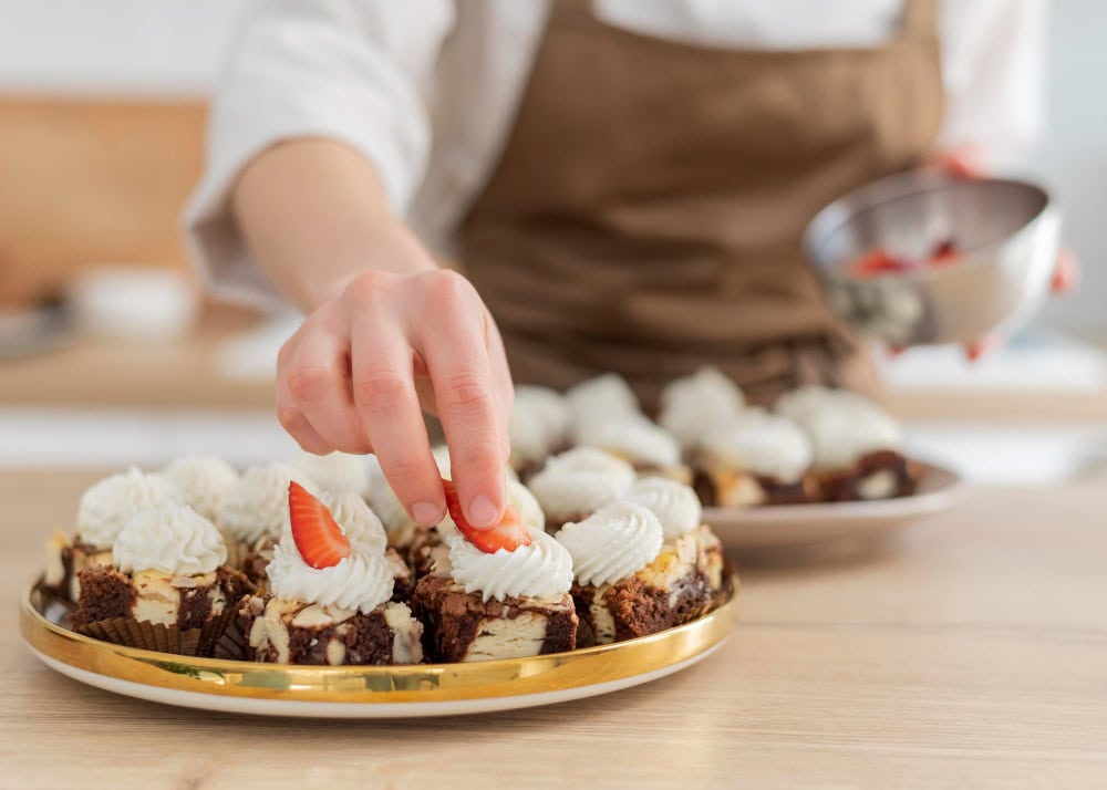 Person in an apron places a strawberry on a platter of bite-size chocolate cakes topped with whipped cream.