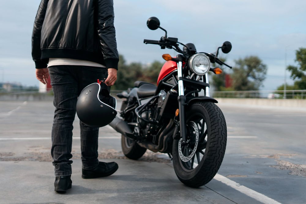 Person in a black jacket and jeans stands beside a red motorcycle, holding a black helmet.