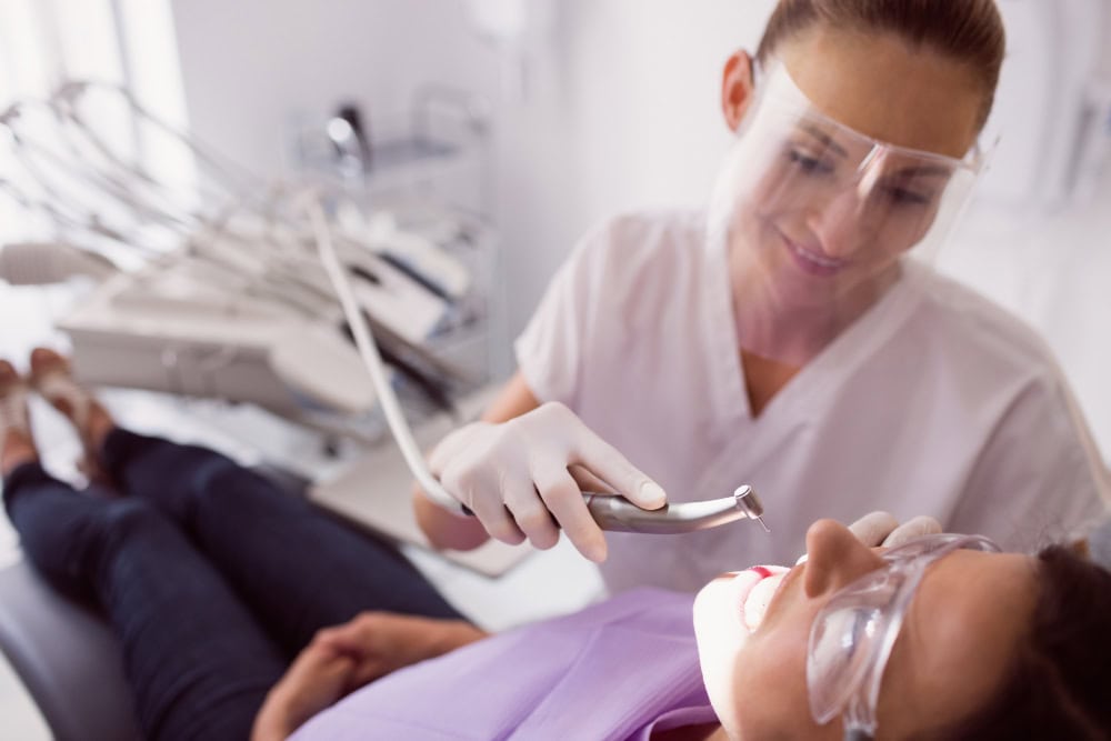 Dentist wearing protective gear drills a patient’s teeth in a dental chair, using a handheld dental drill and suction tube in a clinical setting.
