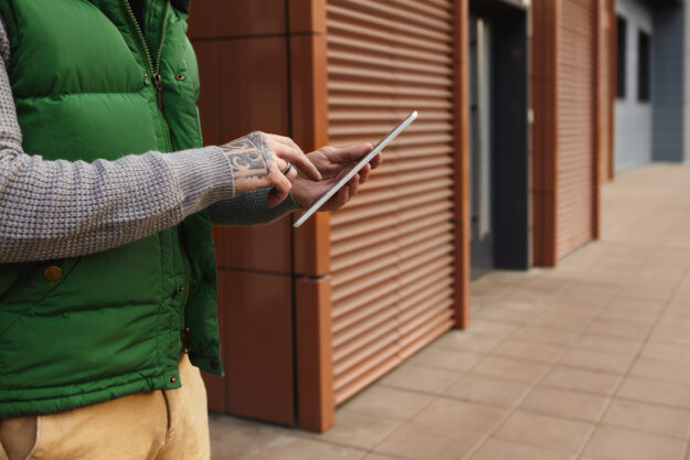 Person in a green puffer vest uses a tablet outdoors beside wooden facade panels, showing a tattooed hand on the device.