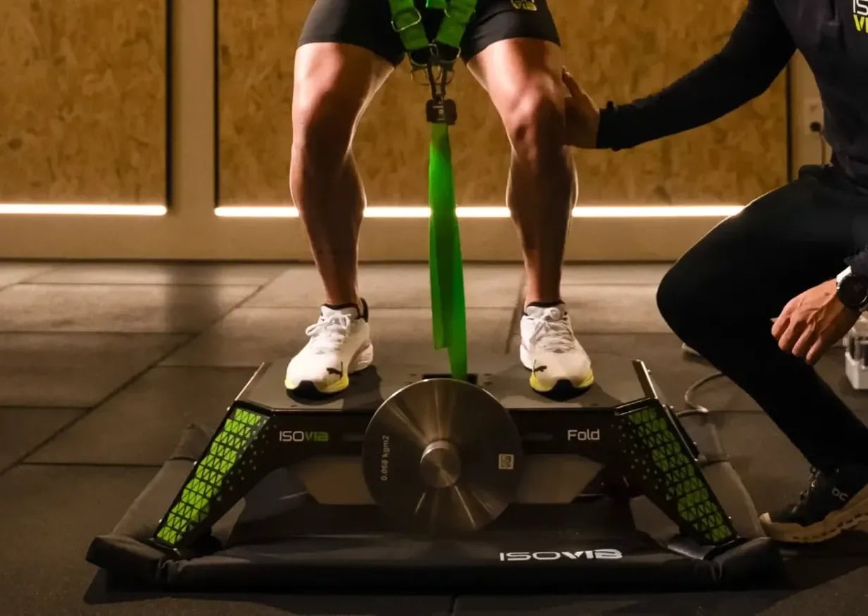 Athlete in sneakers stands on a compact IsoVibe Fold training device with a large central flywheel, trainer beside them.