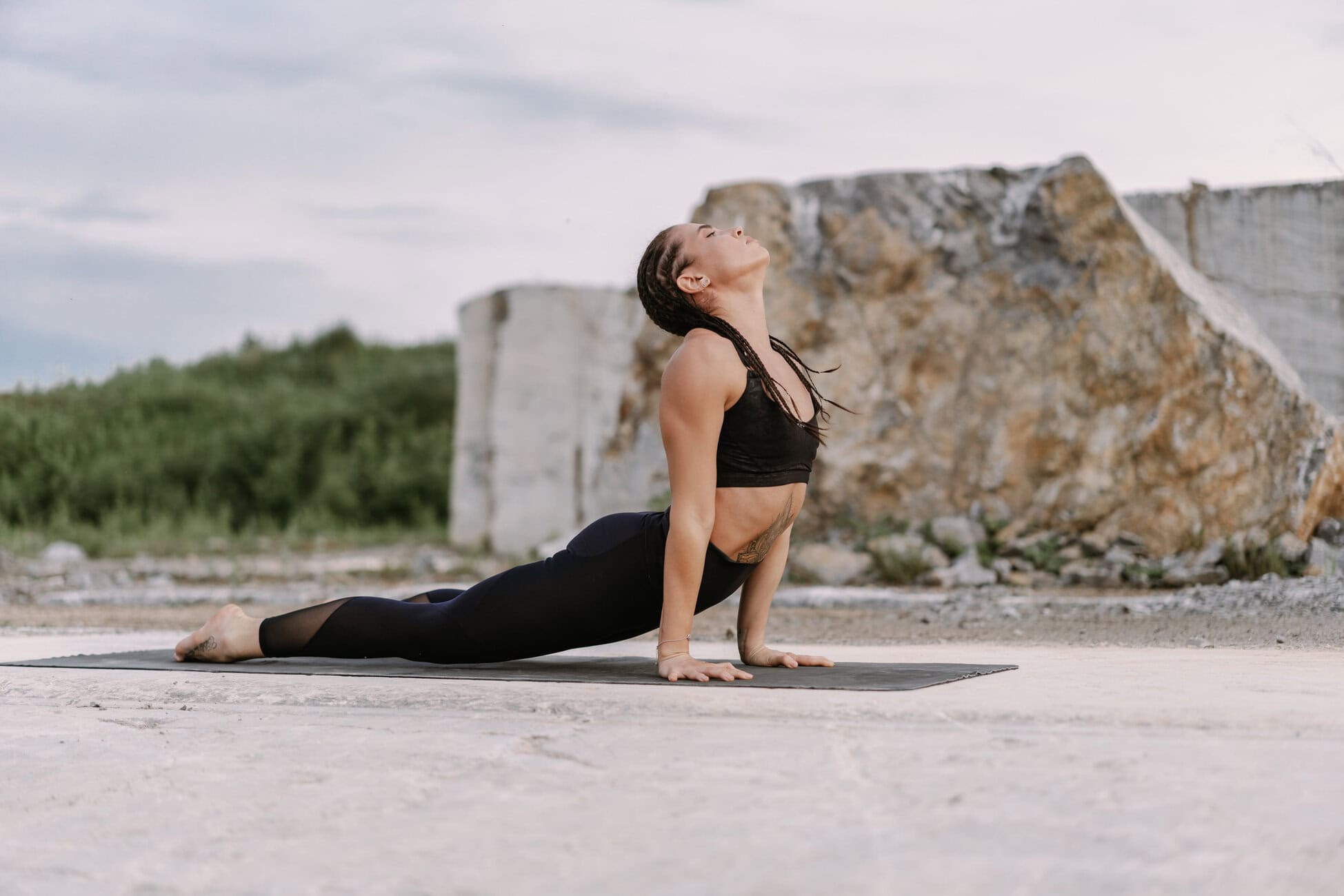 Woman practicing yoga outdoors on a black mat, in a backbend with chest lifted and head tilted back.