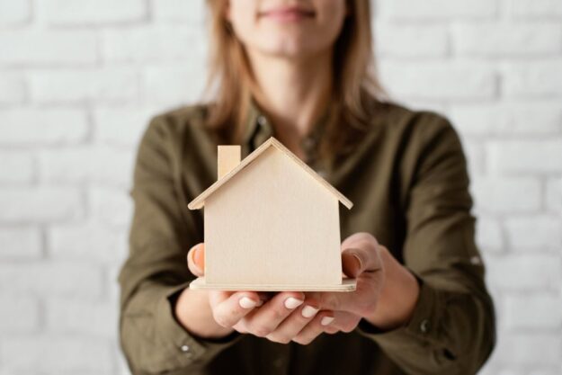 Person presents a small wooden house model toward the camera, symbolizing home ownership or real estate.