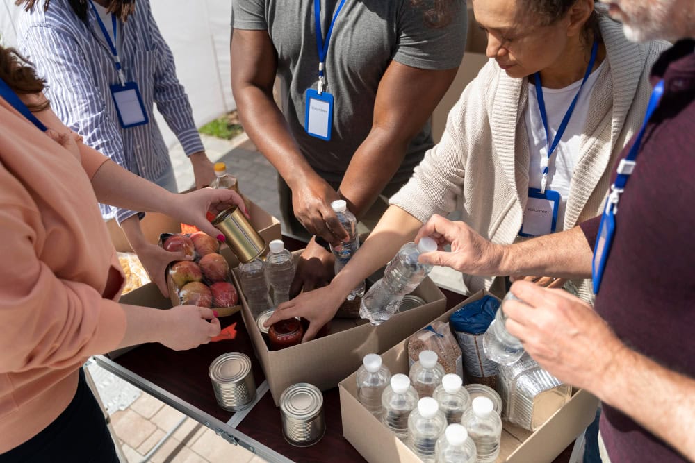Group of volunteers assembling care packages with water bottles, apples, and canned goods on a table.