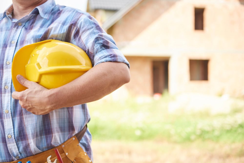 Construction worker in a plaid shirt holding a yellow hard hat at a building site.